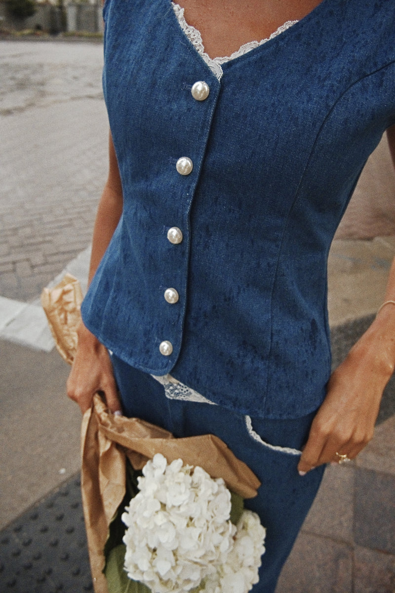 A close-up of a person wearing a blue denim vest with silver buttons and white lace trim, holding a brown paper bag with white flowers, outdoors on a paved street.