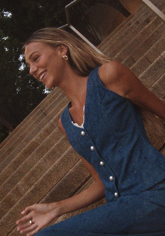A woman wearing a blue denim sleeveless jumpsuit with pearl buttons, sitting on outdoor stone steps.