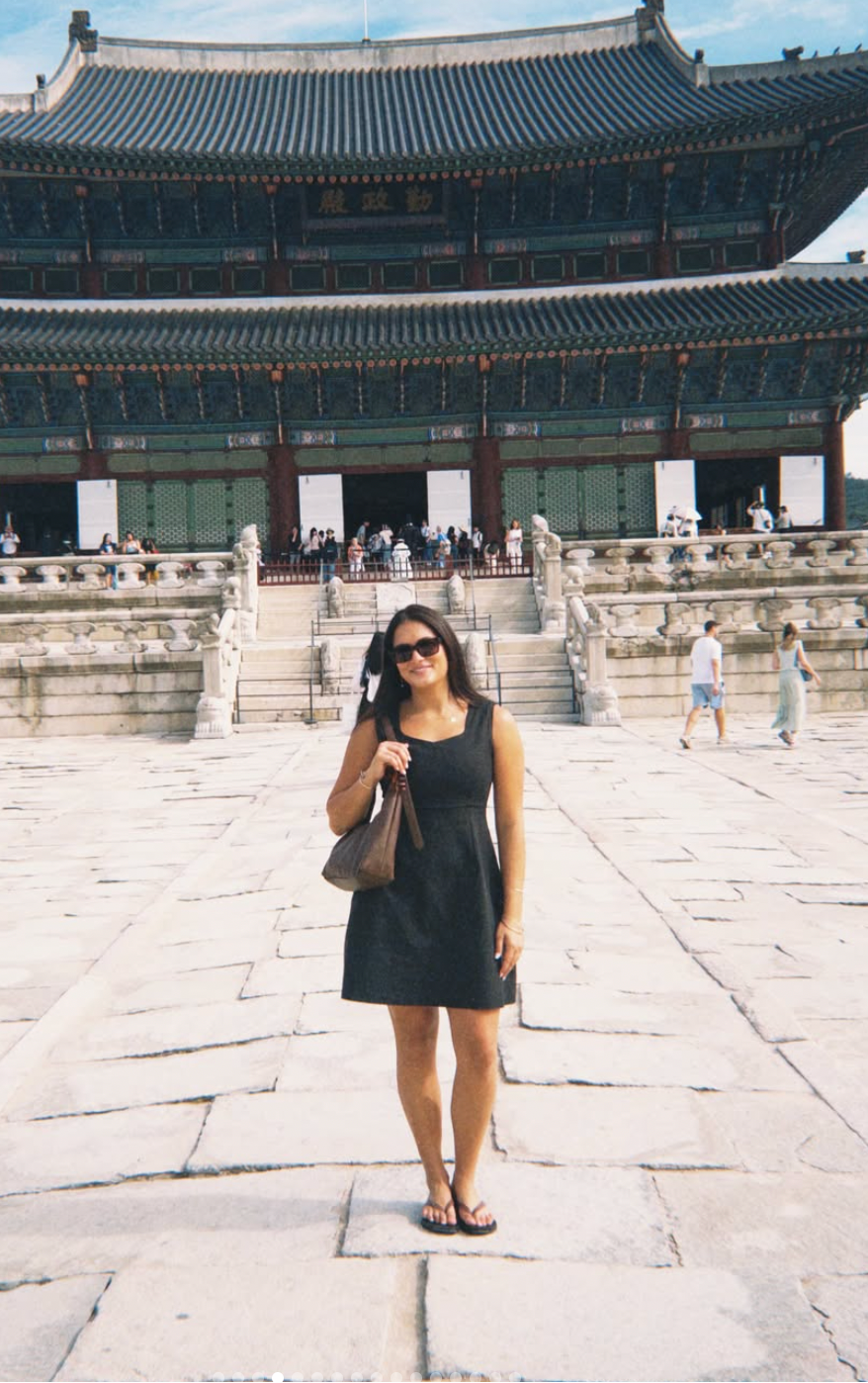 A woman wearing a black sleeveless dress and sunglasses stands in a stone courtyard in front of a large traditional Korean palace building.