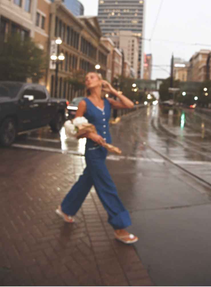 A woman in a blue cotton denim sleeveless top and matching wide-leg pants walks across a wet city street at dusk, holding a white bouquet. 